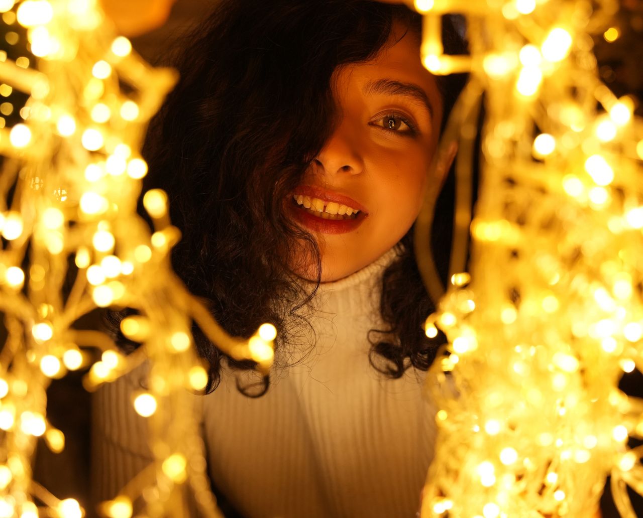 A young woman smiling through golden string lights, creating a warm and festive ambiance.