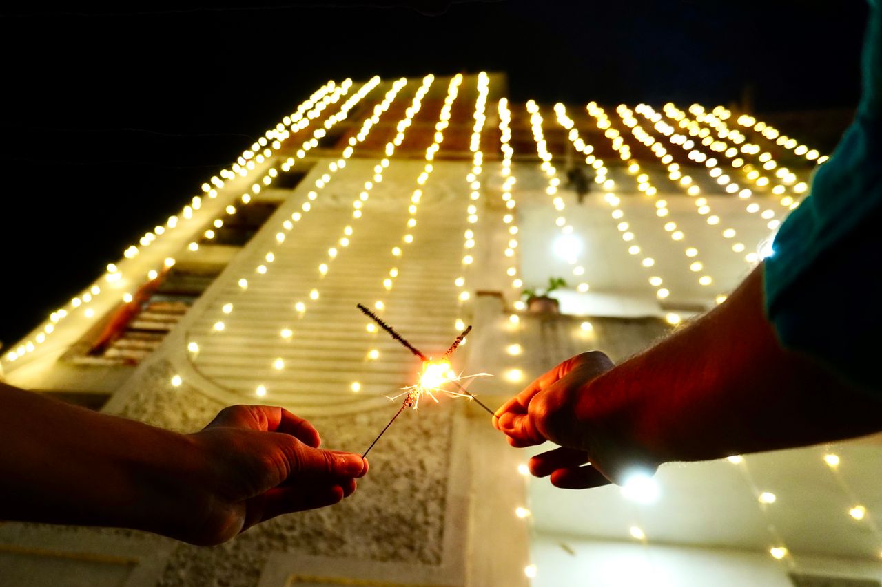 Hands holding sparklers in front of a brightly lit building during Diwali festivities at night.