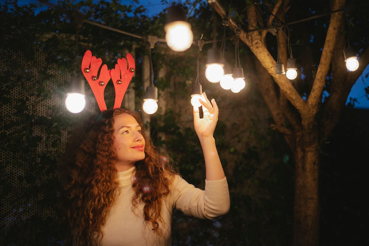 Smiling adult female in decorative deer horns enjoying glowing garland lights during New Year holiday at night