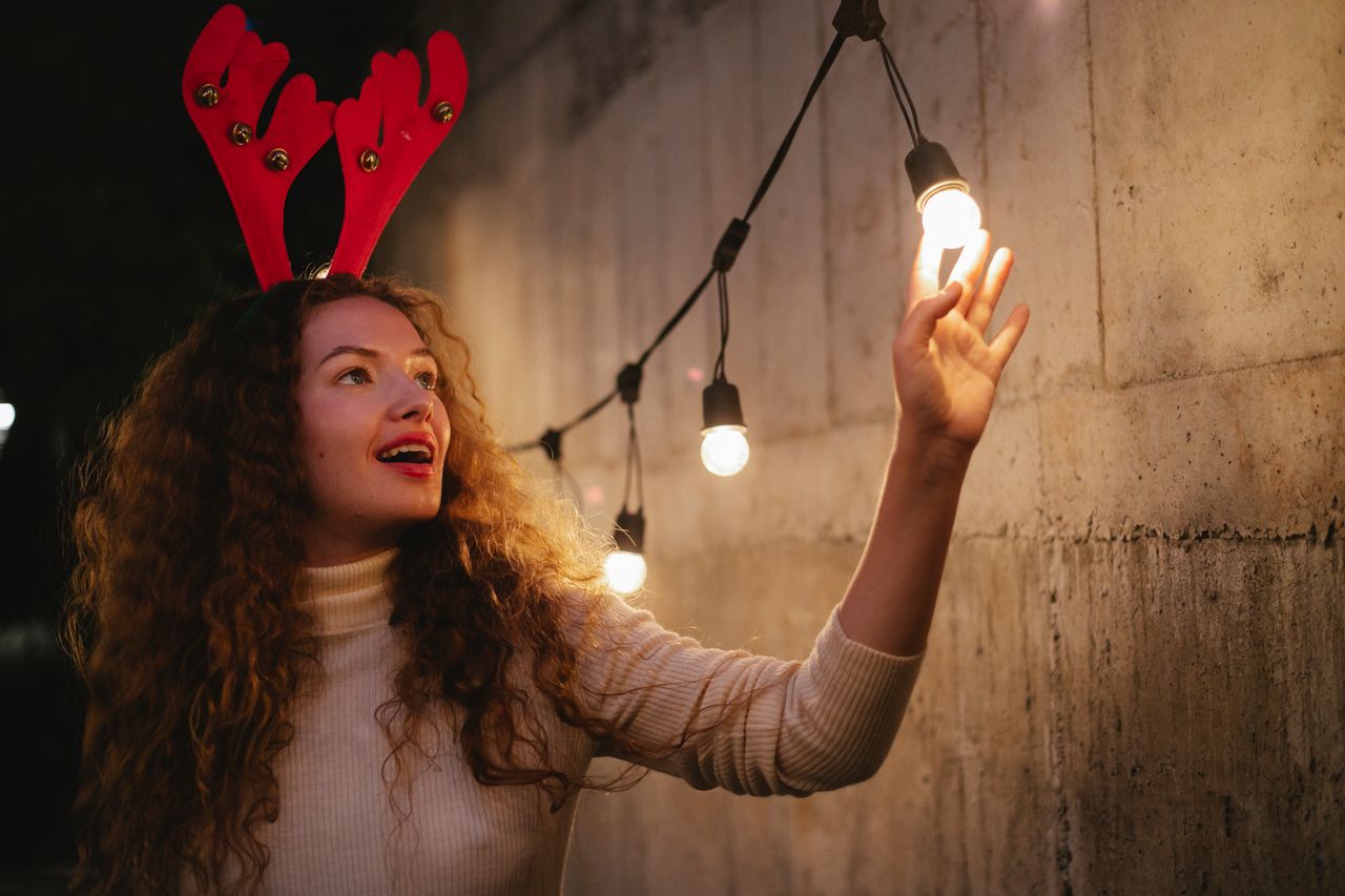 Happy female with curly hair in deer headband touching shining light bulb of garland decorating concrete wall for Christmas